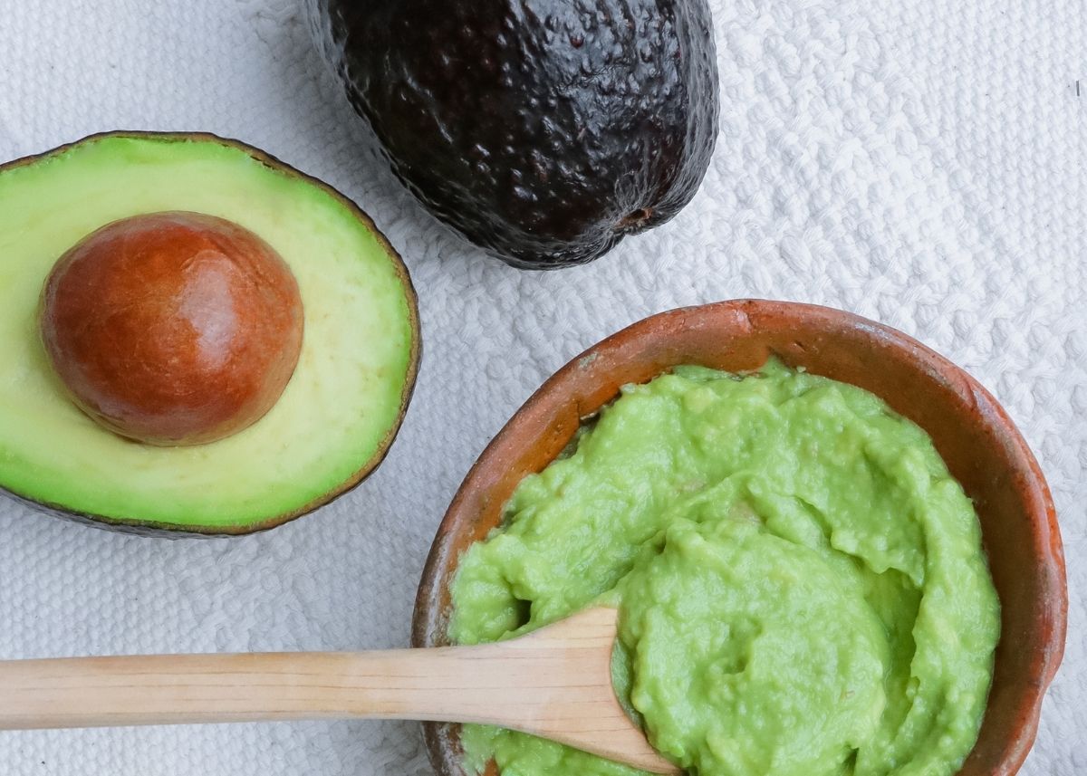 Sliced avocado next to small brown bowl of mashed avocado and wooden spoon.