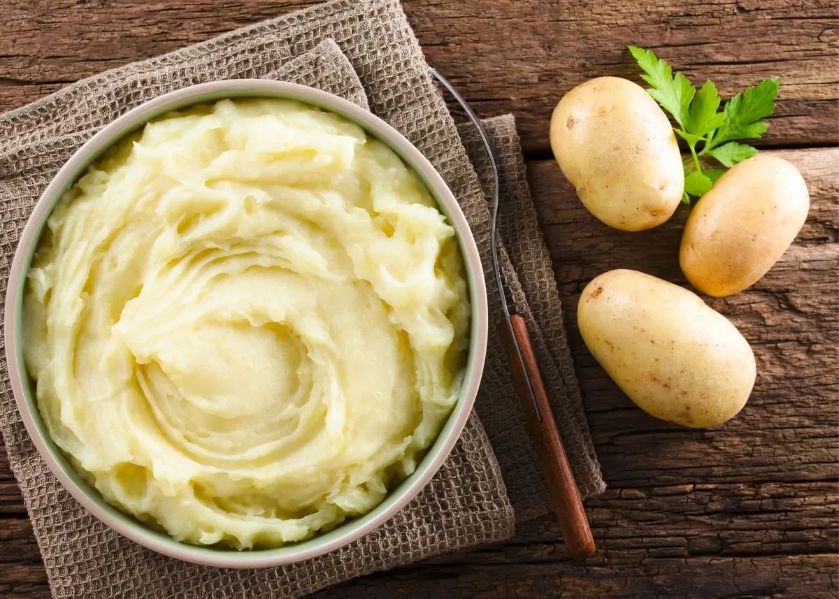 Large bowl filled with mashed potatoes next to three whole potatoes on table.