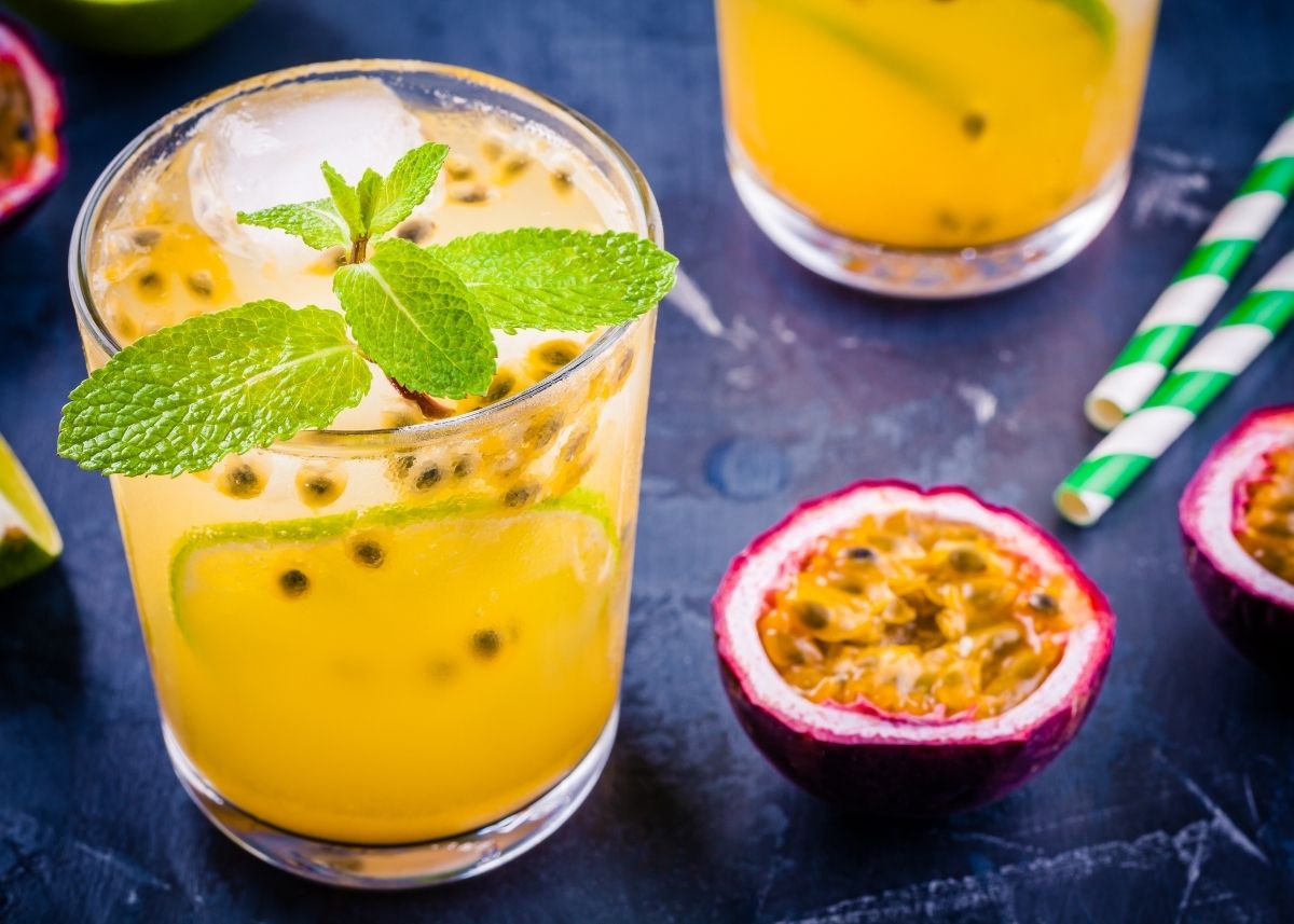 Half of a passion fruit next to two glasses of passion fruit juice with sprig of garnish.