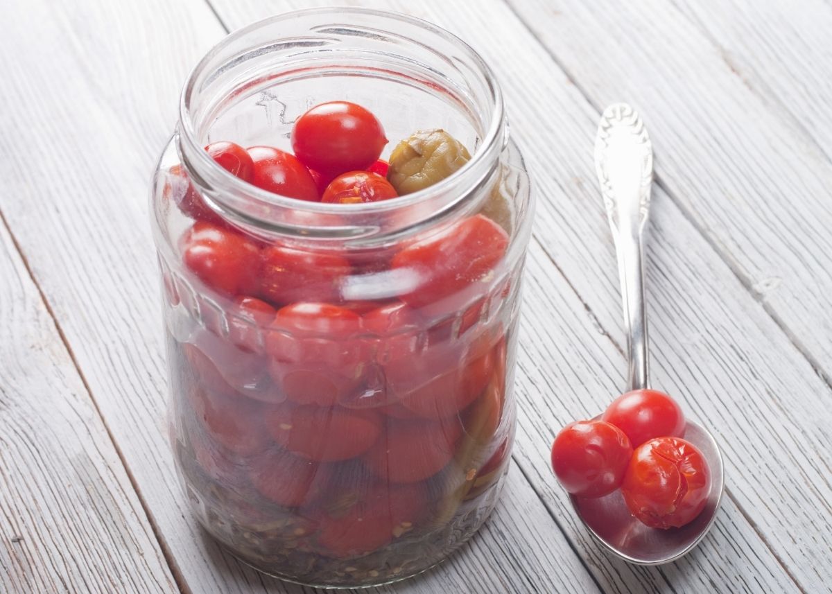 Clear glass mason jar filled with pickled cherry tomatoes next to a metal spoon.