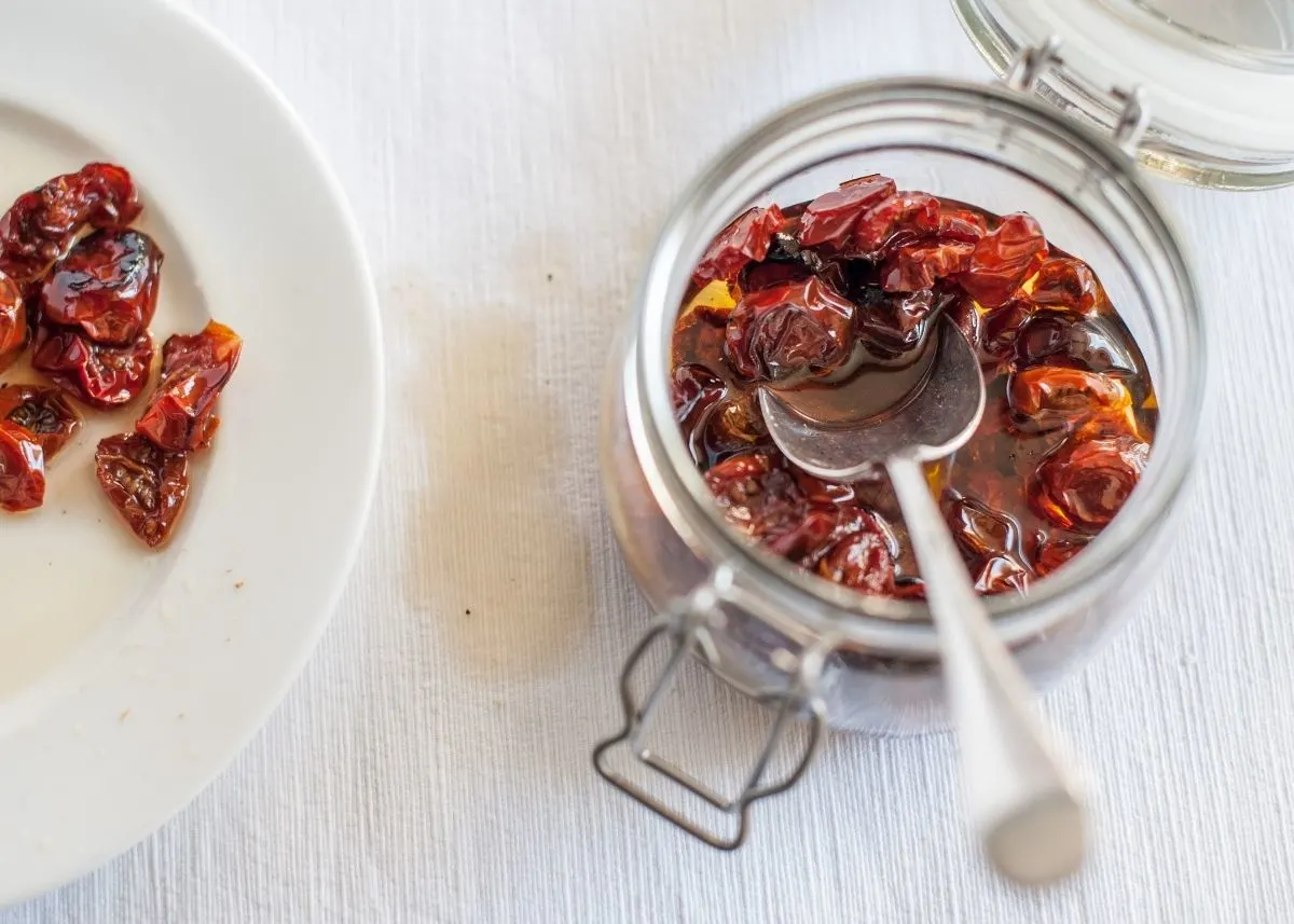 Clear glass jar filled with sundried tomatoes in oil and a metal spoon.