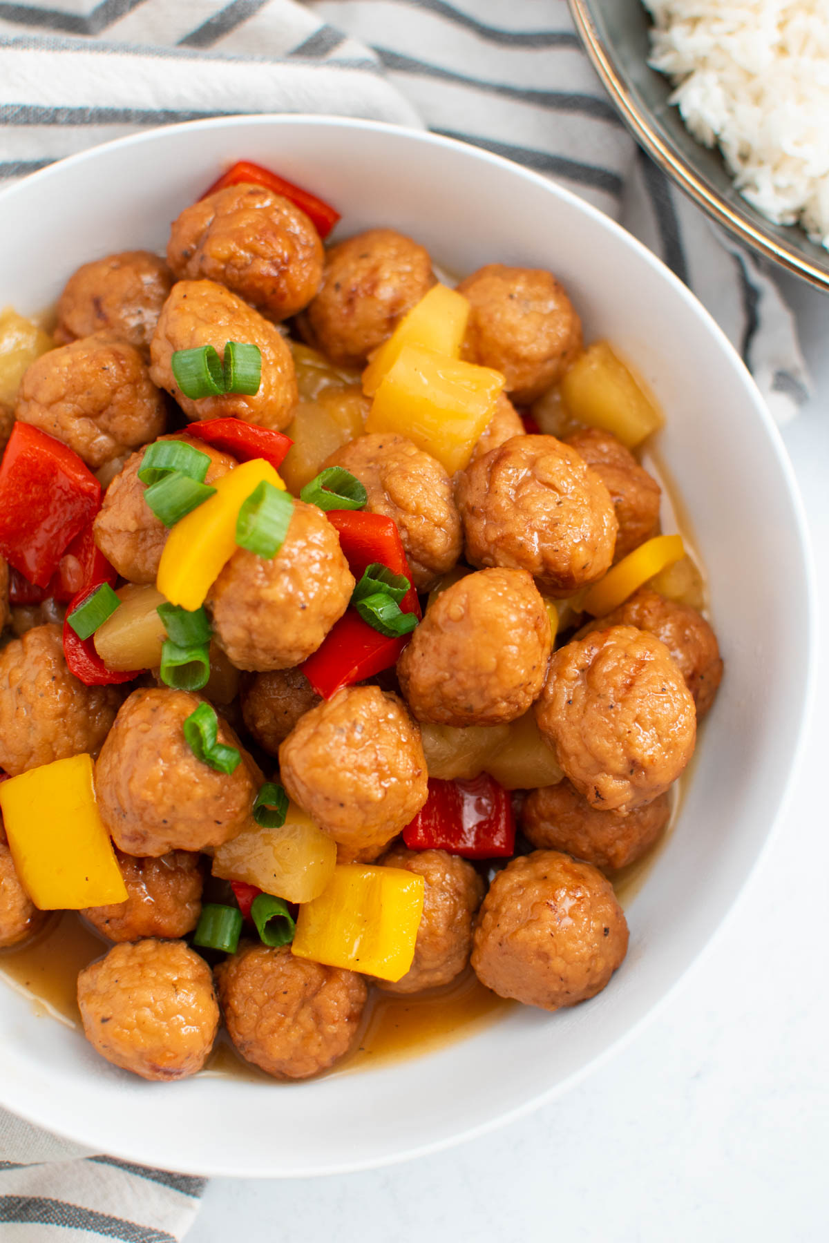 Sweet and sour meatballs in white bowl with pineapple chunks and chopped green onion garnish.
