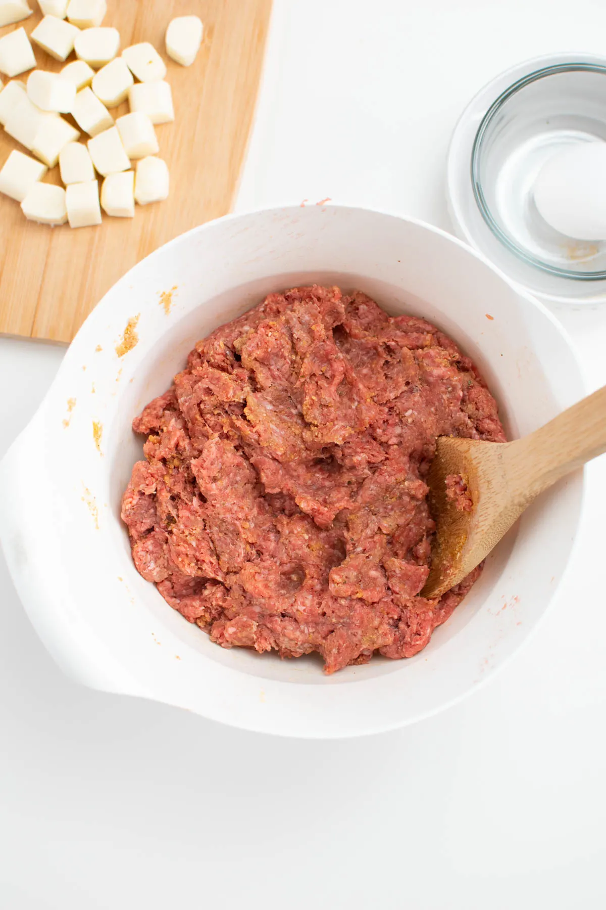 Raw hamburger meat mixture in white bowl with wooden spoon.
