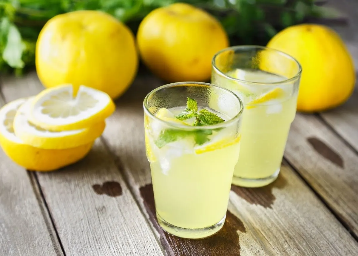 Two glasses of lemon juice on a wooden table surrounded by whole and sliced lemons.