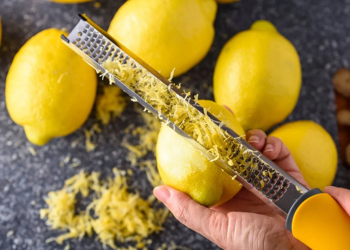 Woman uses a microplane grater to zest a lemon over a table with lemons on it.