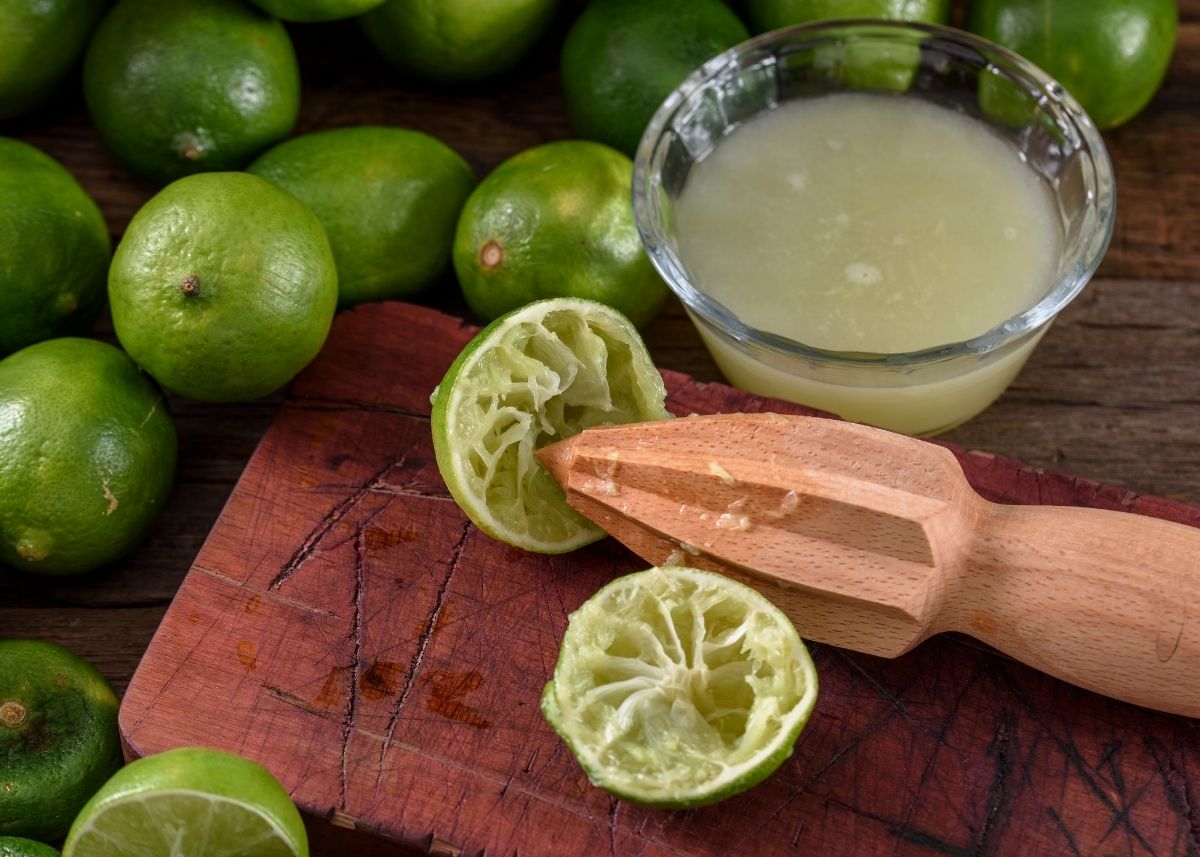 Whole limes surround a wooden cutting board with two juiced limes and a bowl of lime juice.