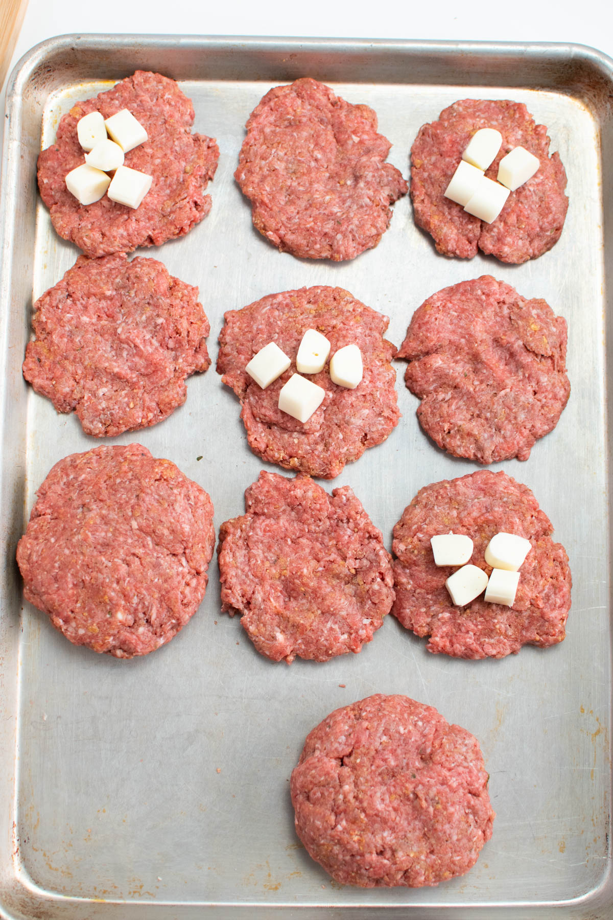 Mozzarella cubes on some raw hamburger patties all on sheet pan.