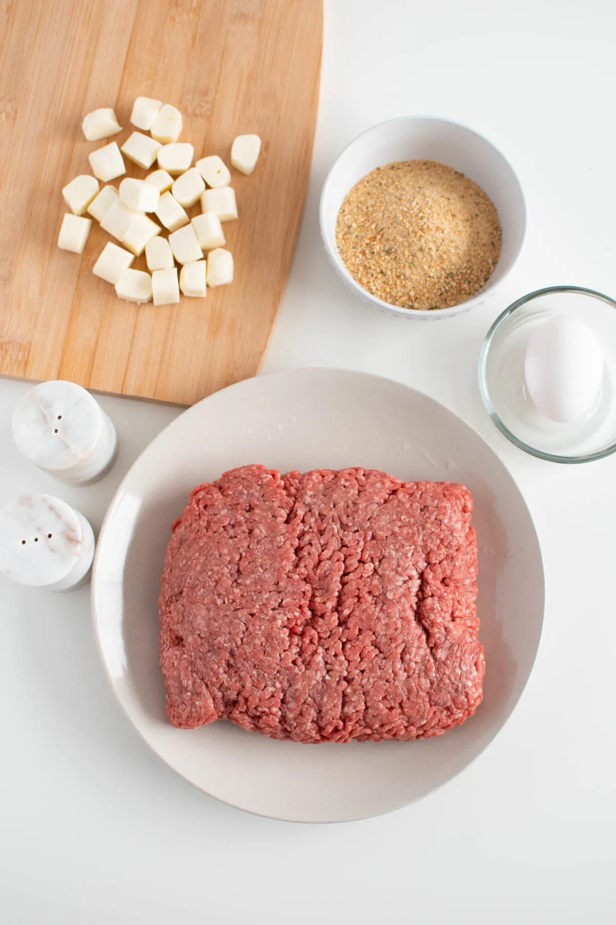 Mozzarella stuffed burger ingredients on table including ground beef, mozzarella cheese cubes, and bread crumbs.