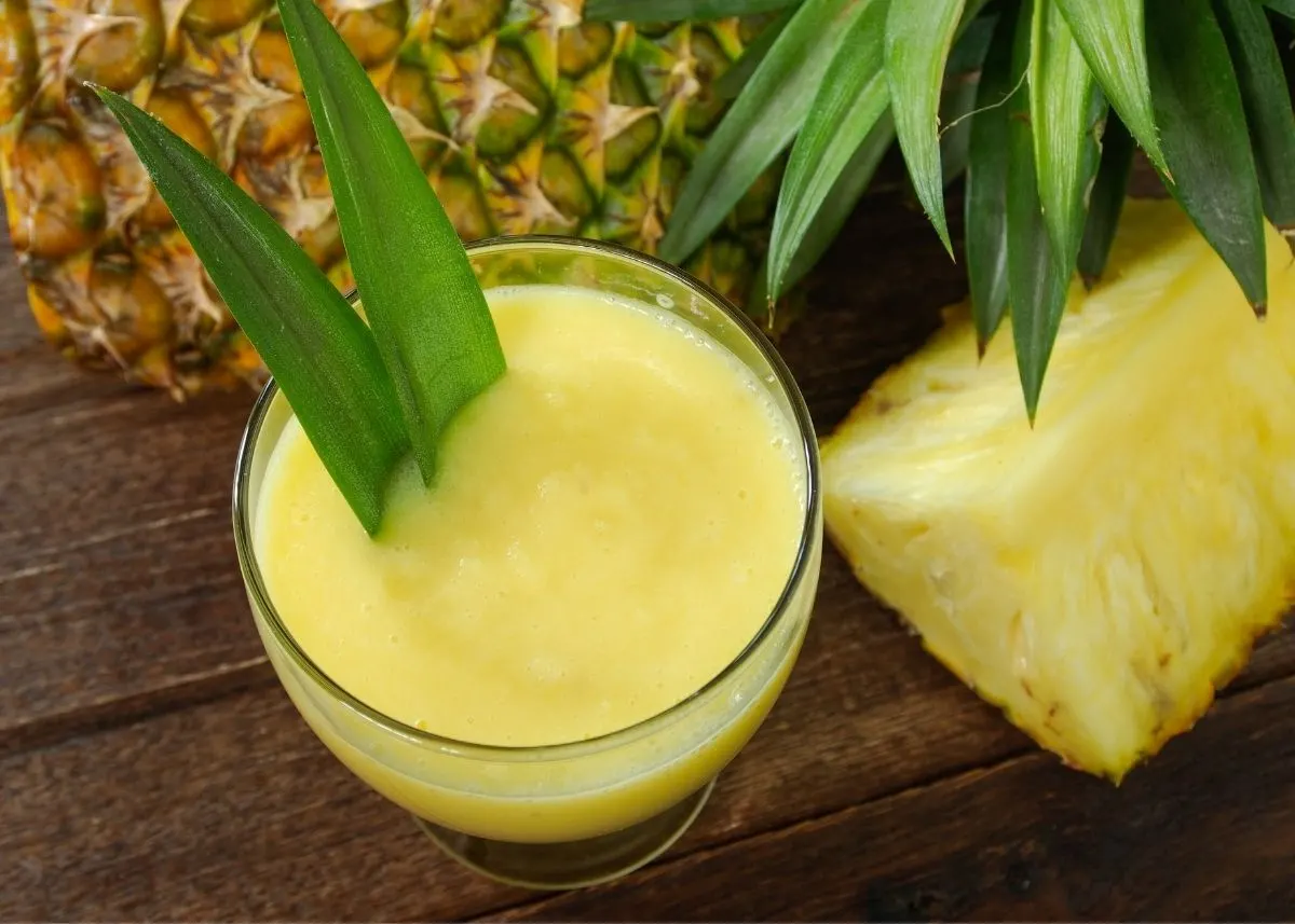 Glass of pineapple juice with green leaf garnish next to whole and cut pineapple.