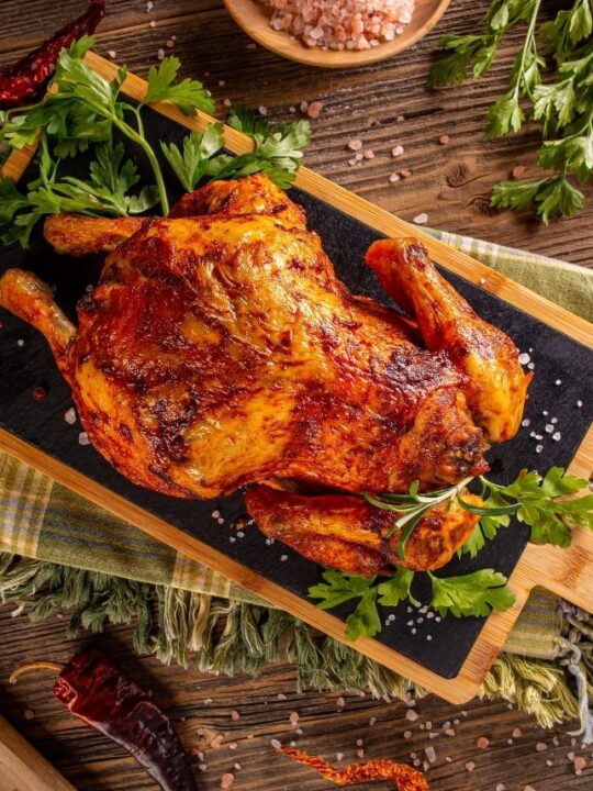 Crispy whole rotisserie chicken on a platter surrounded by green garnish on wooden table.