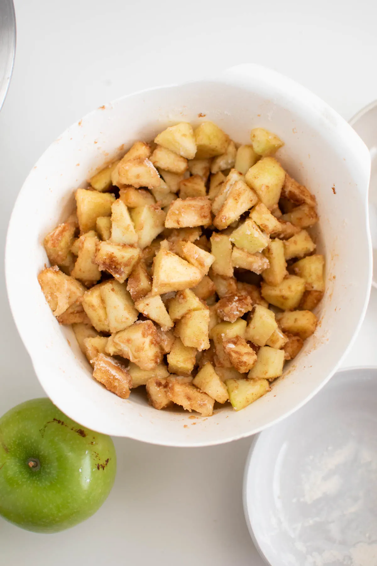 Cinnamon apple pieces in white mixing bowl surrounded by Granny Smith apple.