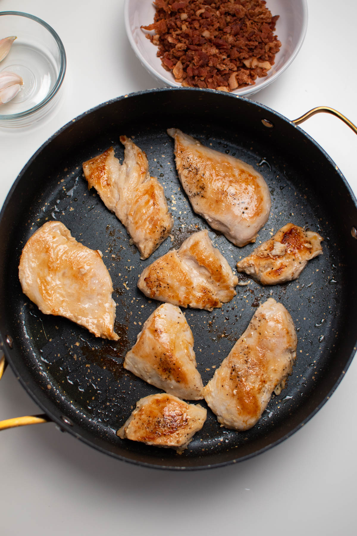 Several cooked chicken pieces in black skillet with bowls of other ingredients nearby.