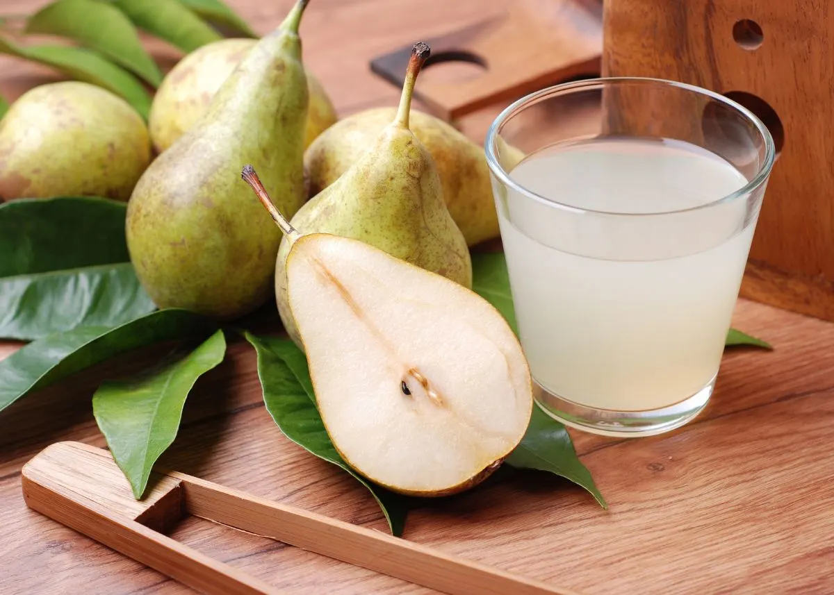 Whole and sliced pears sitting on leaves next to a glass of pear juice.