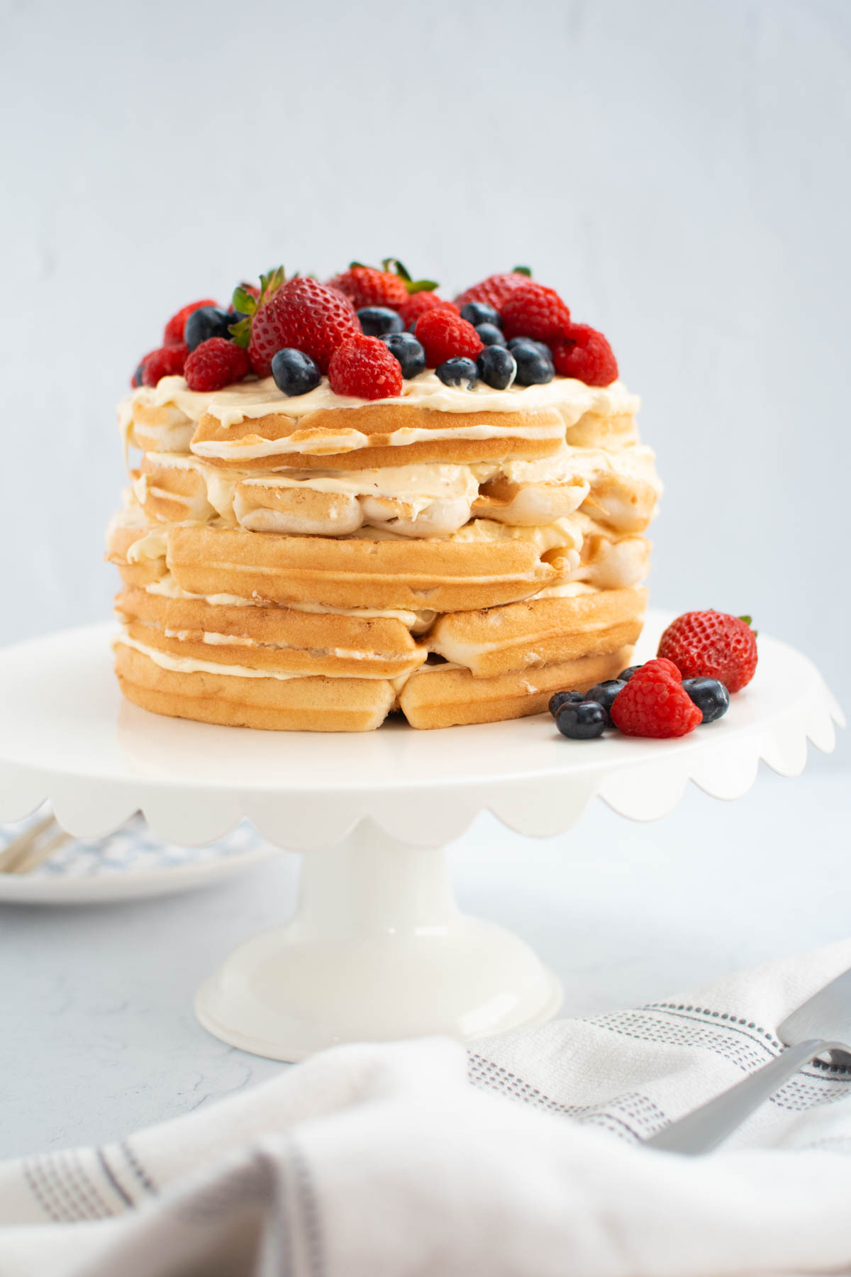 Five layer berry waffle cake on white cake stand with white towel nearby.