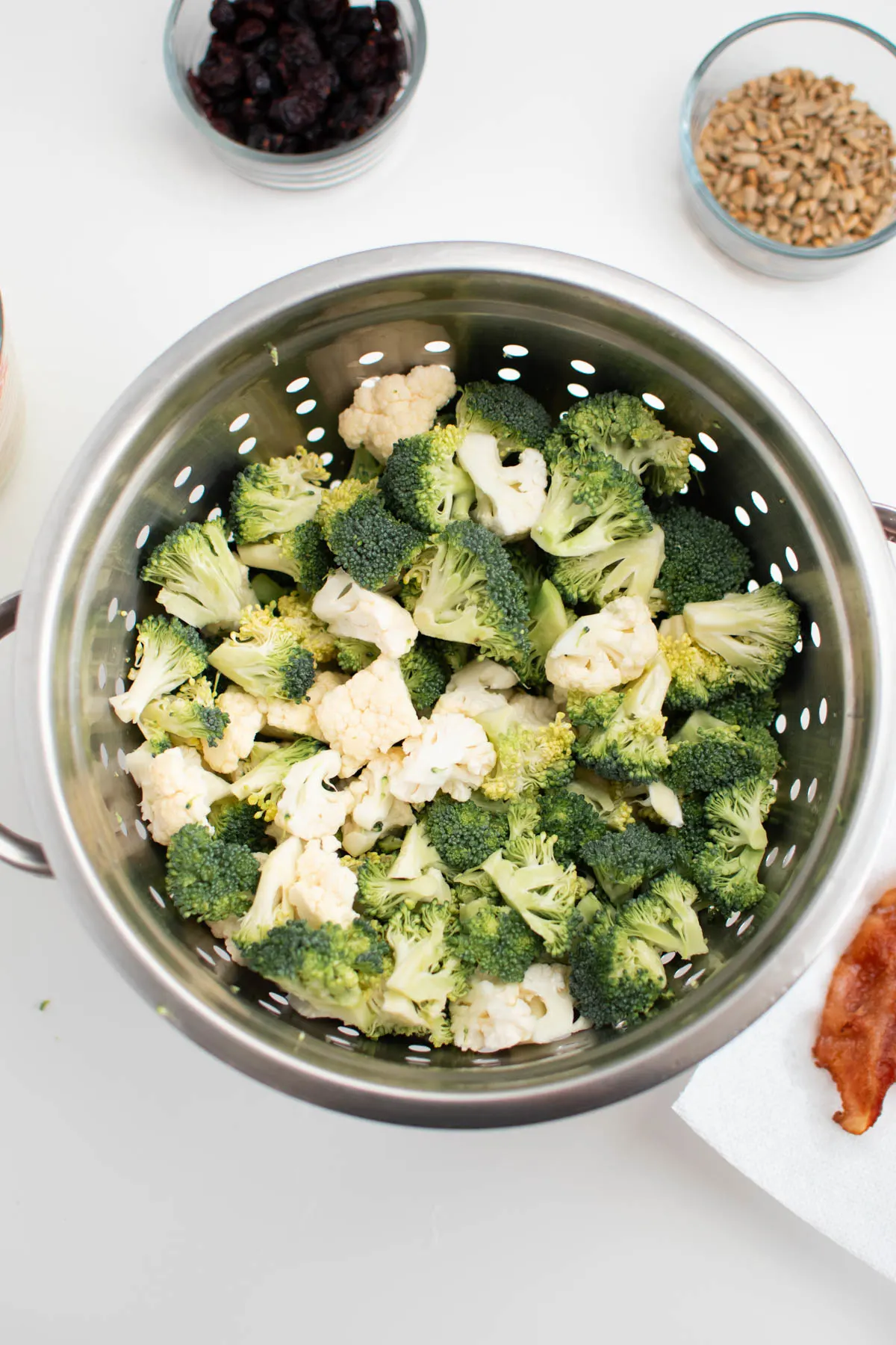 Broccoli and cauliflower florets in strainer on white table with bowls of ingredients nearby.