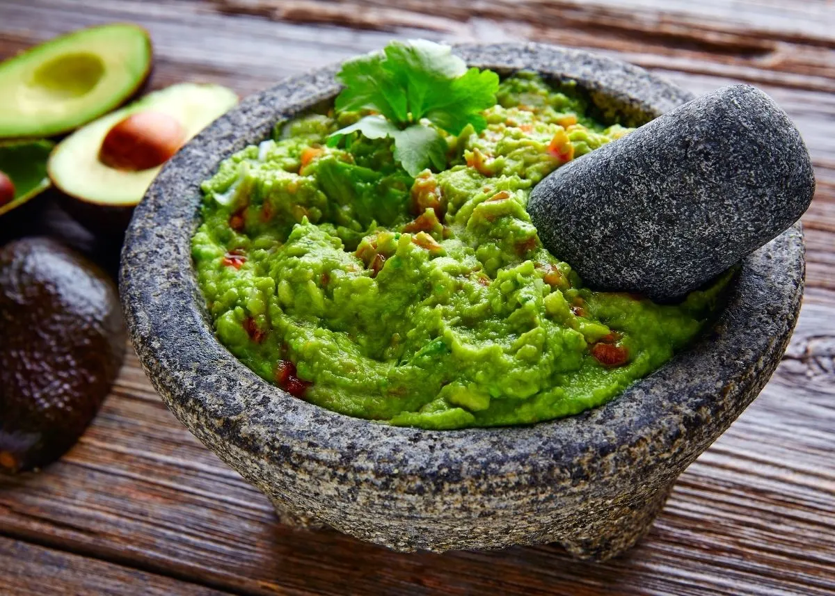 Homemade guacamole in a mortar and pestle with herbs next to whole and cut avocados.