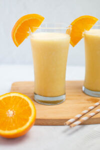 Two glasses of Orange Julius with orange segments on rim and orange half nearby.