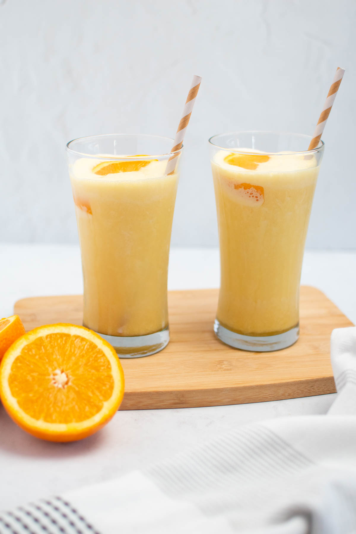 Two glasses of Orange Julius with striped straws and orange segments, all on wood cutting board.