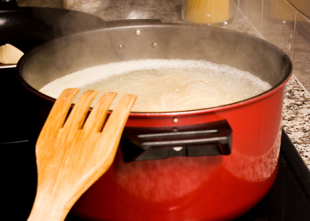 Large red cook pot filled with simmering pasta water with a wooden kitchen utensil.