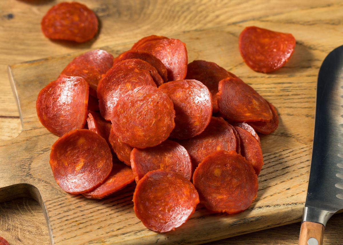 Several slices of dark red pepperoni piled on a light wood cutting board.
