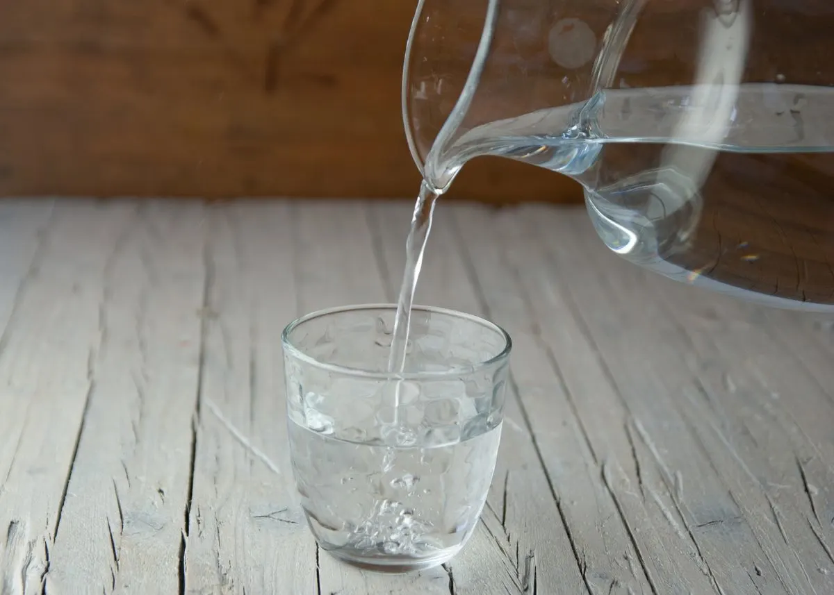 Glass pitcher of water pouring water into a small drinking glass on a wooden table.