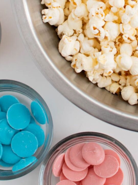 Orange, blue, and pink candy melts in kitchen prep bowls next to a bowl of popcorn.
