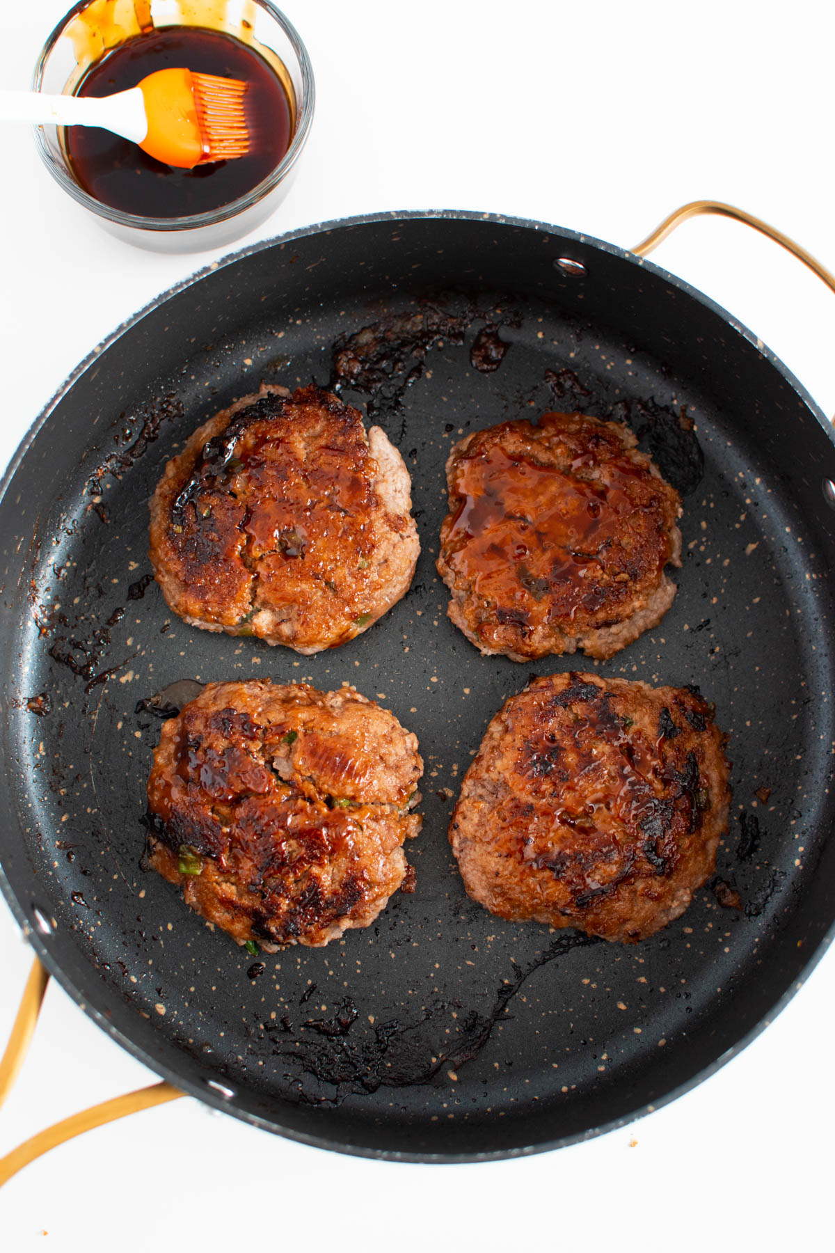 Four cooked turkey burgers basted with teriyaki sauce in black frying pan.