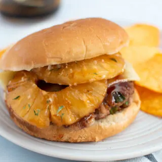 Hawaiian turkey burger with pineapple slices on plate with potato chips and teriyaki sauce in background.