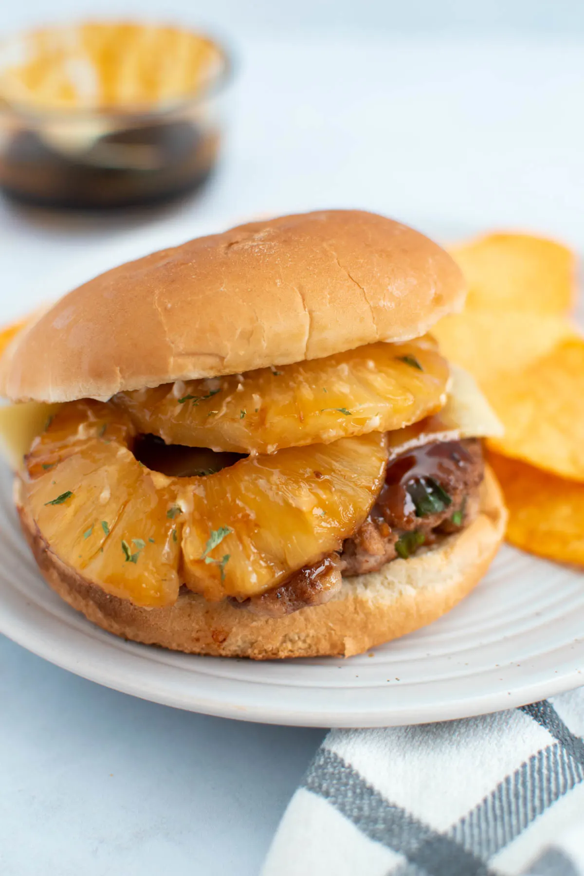 Hawaiian turkey burger with pineapple slices on plate with potato chips and teriyaki sauce in background.