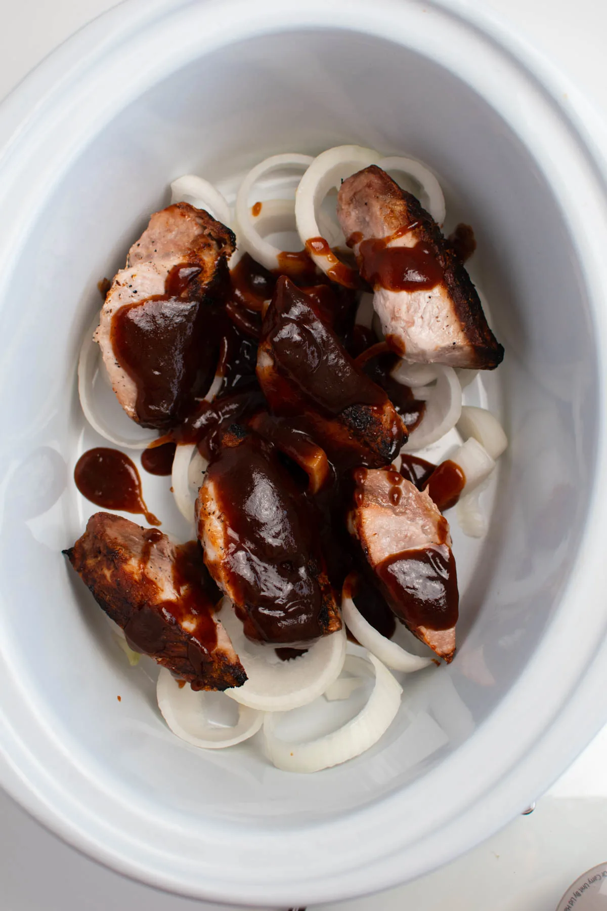 Barbeque sauce on ribs and onion slices in white Crock Pot insert.