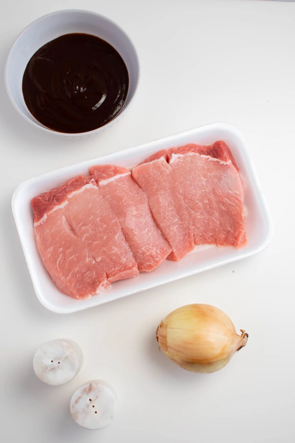 Country style rib ingredients on white table including raw ribs, onion, and barbeque sauce.