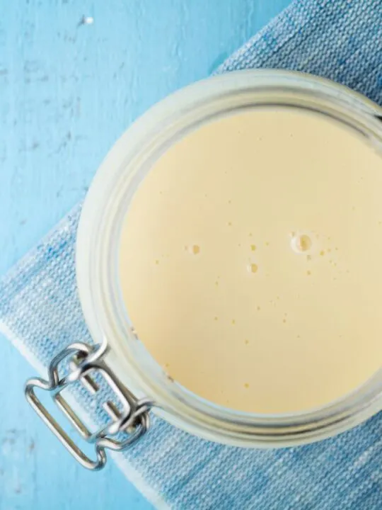 Evaporated milk in a clear glass canister on a blue background with spoon.