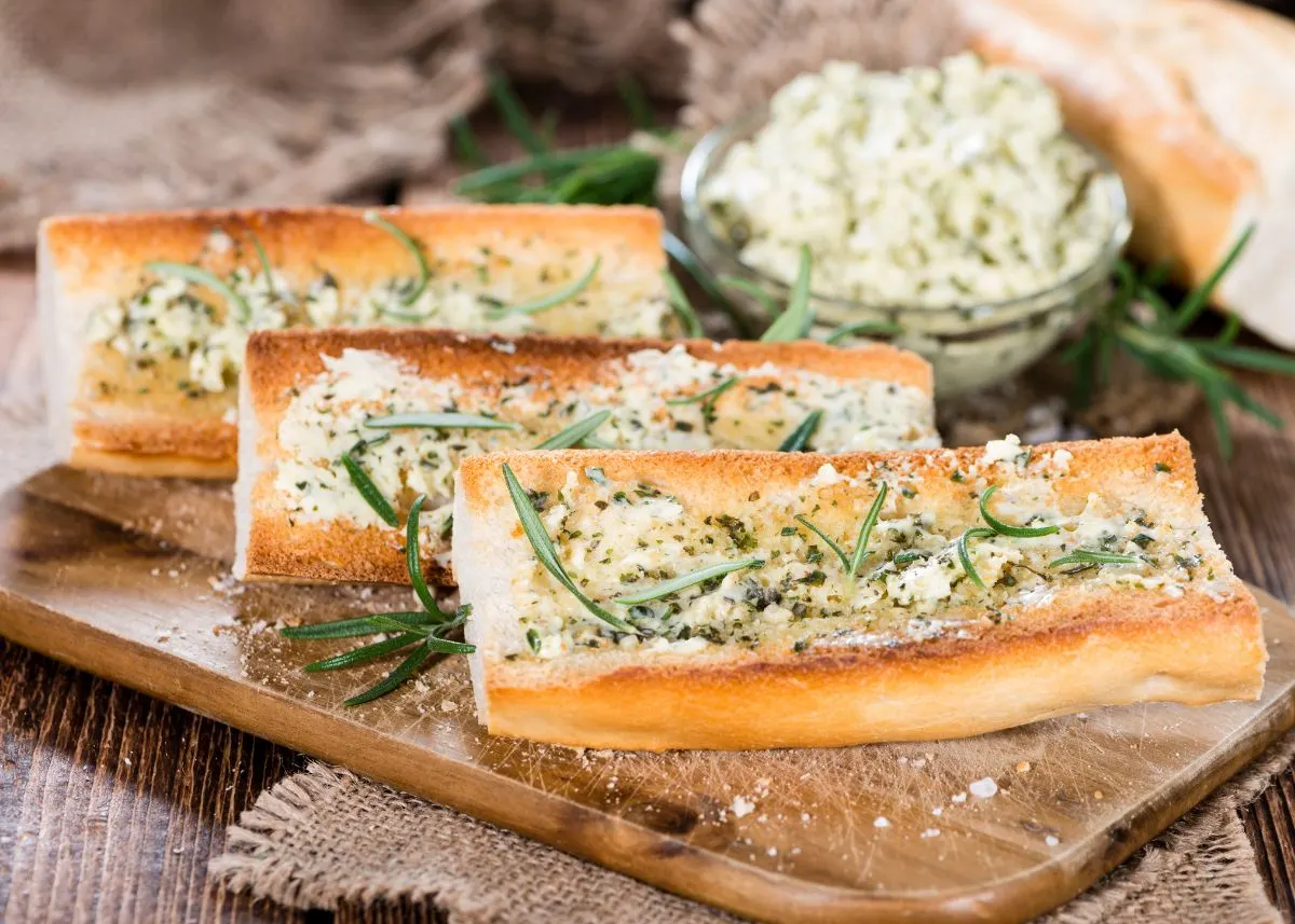Three loaves of garlic bread on a wooden cutting board with fresh herbs and cheese on top.