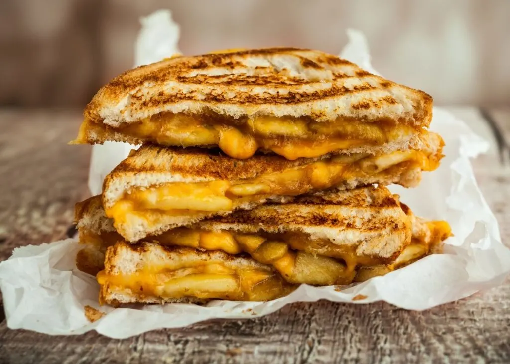 Stack of three grilled cheese sandwiches on a white cloth and brown kitchen table.