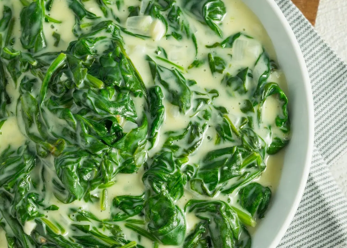 Close up of a large white bowl filled with creamed spinach next to a kitchen towel.