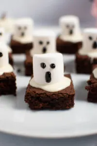 Ghost brownies with marshmallows and frosting gel faces on white platter.