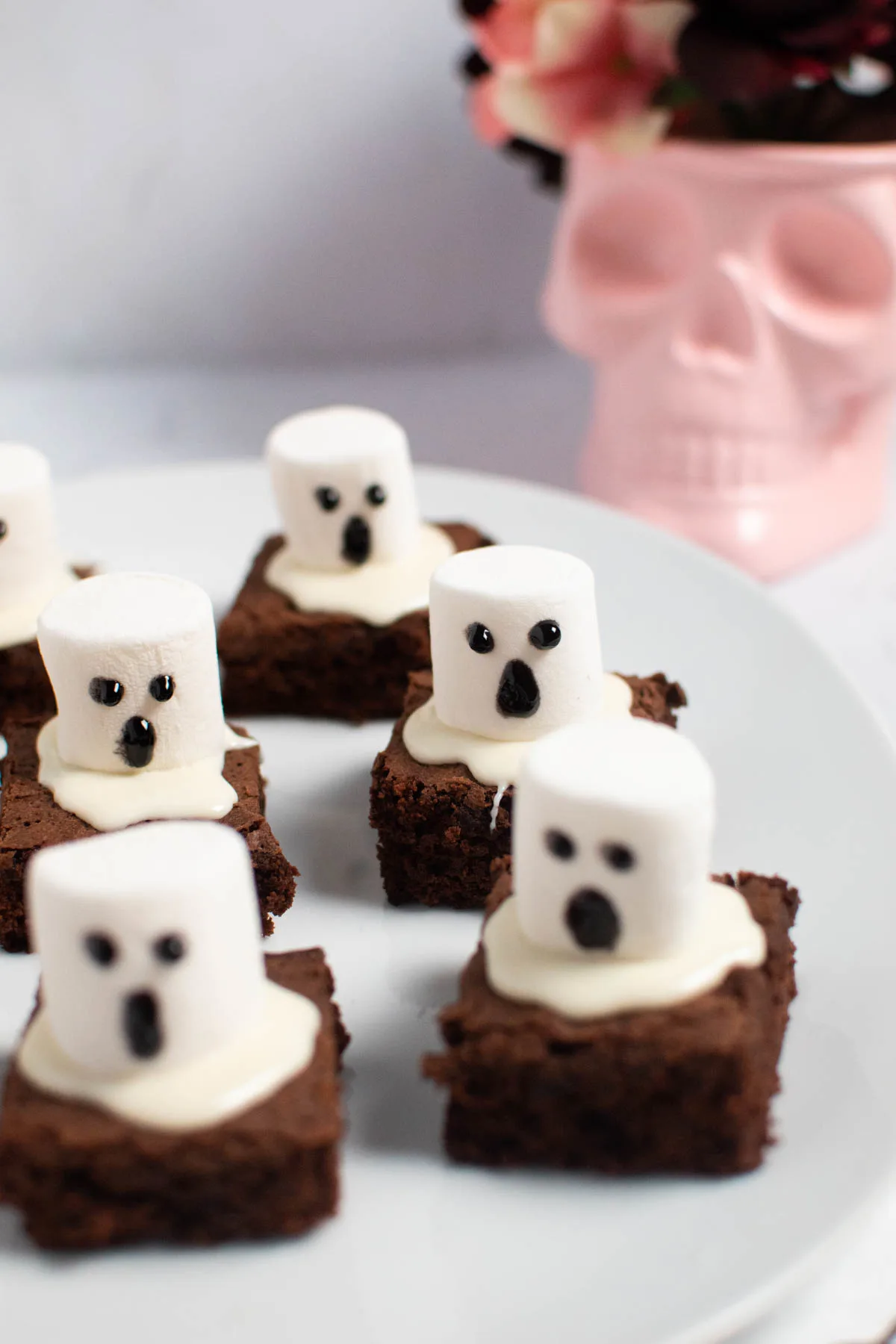 Ghost marshmallow brownies on white platter with pink skull vase in background.