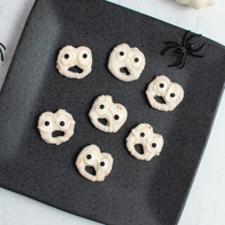 Seven white chocolate ghost pretzels on gray square plate with plastic spiders nearby.