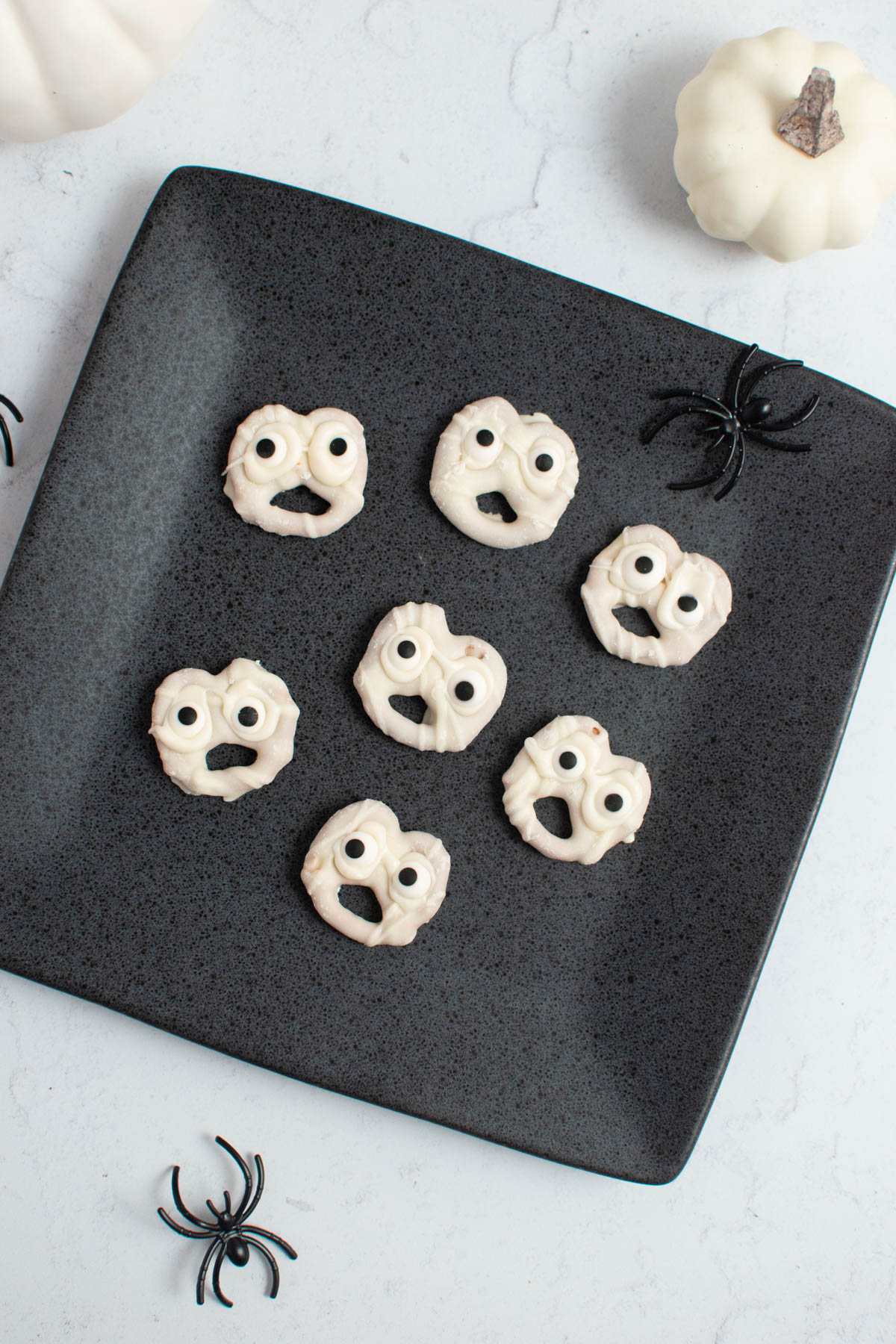 Seven white chocolate ghost pretzels on gray square plate with plastic spiders nearby.