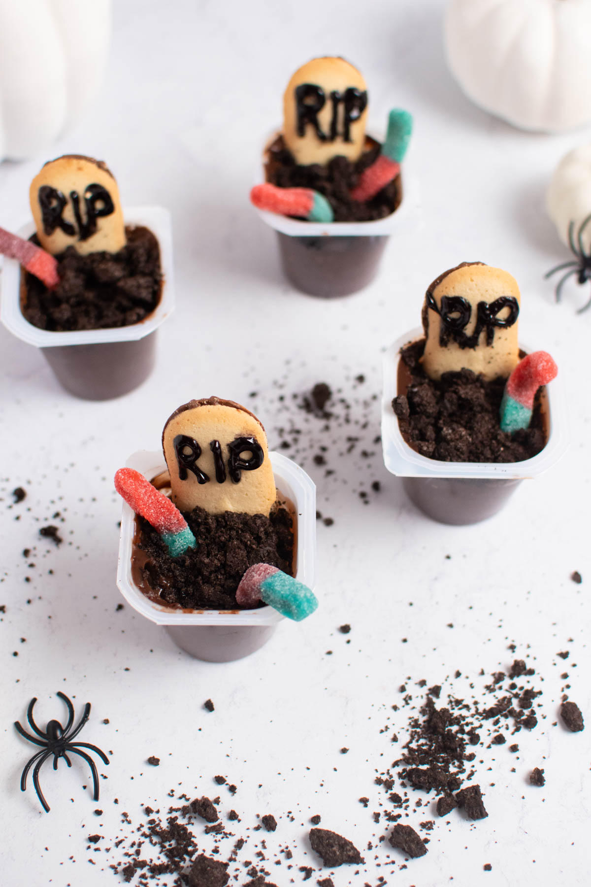 Four graveyard pudding cups on white table with cookie crumbs, plastic spiders, and white pumpkins.