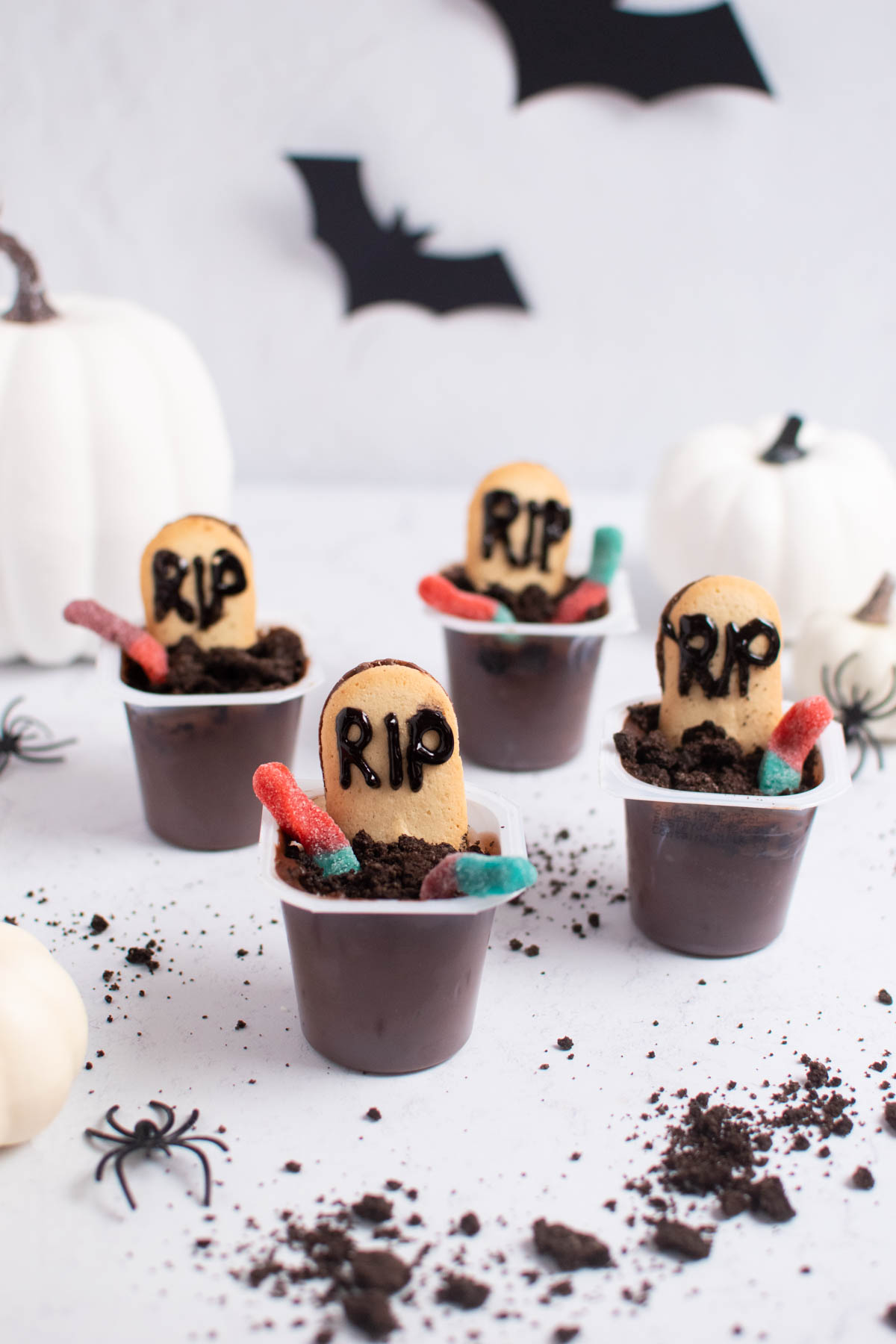 Four Halloween dirt cups with cookies and gummy worms on white table with paper bats in background.
