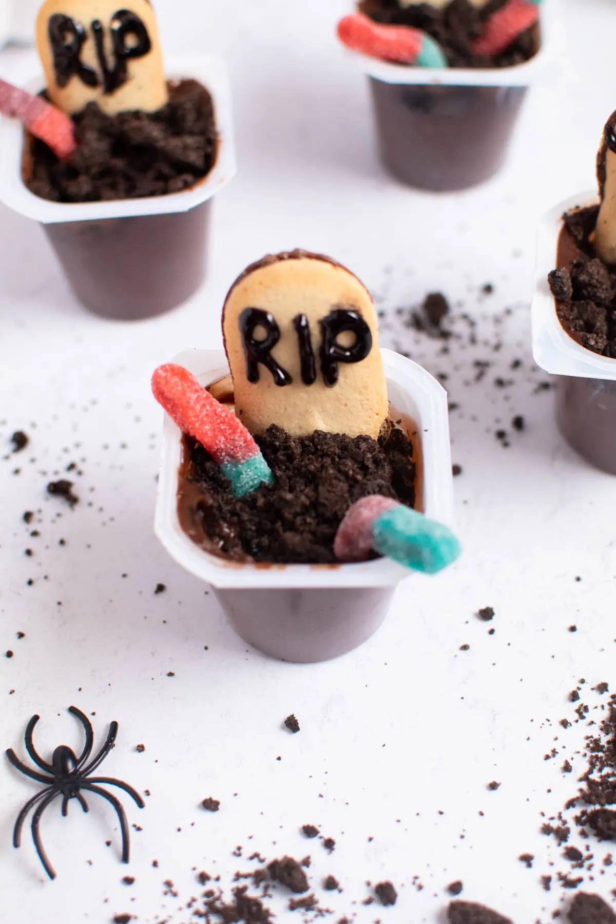 Halloween dirt pudding cups with gummy worms on white table with cookie crumbs and plastic spiders.