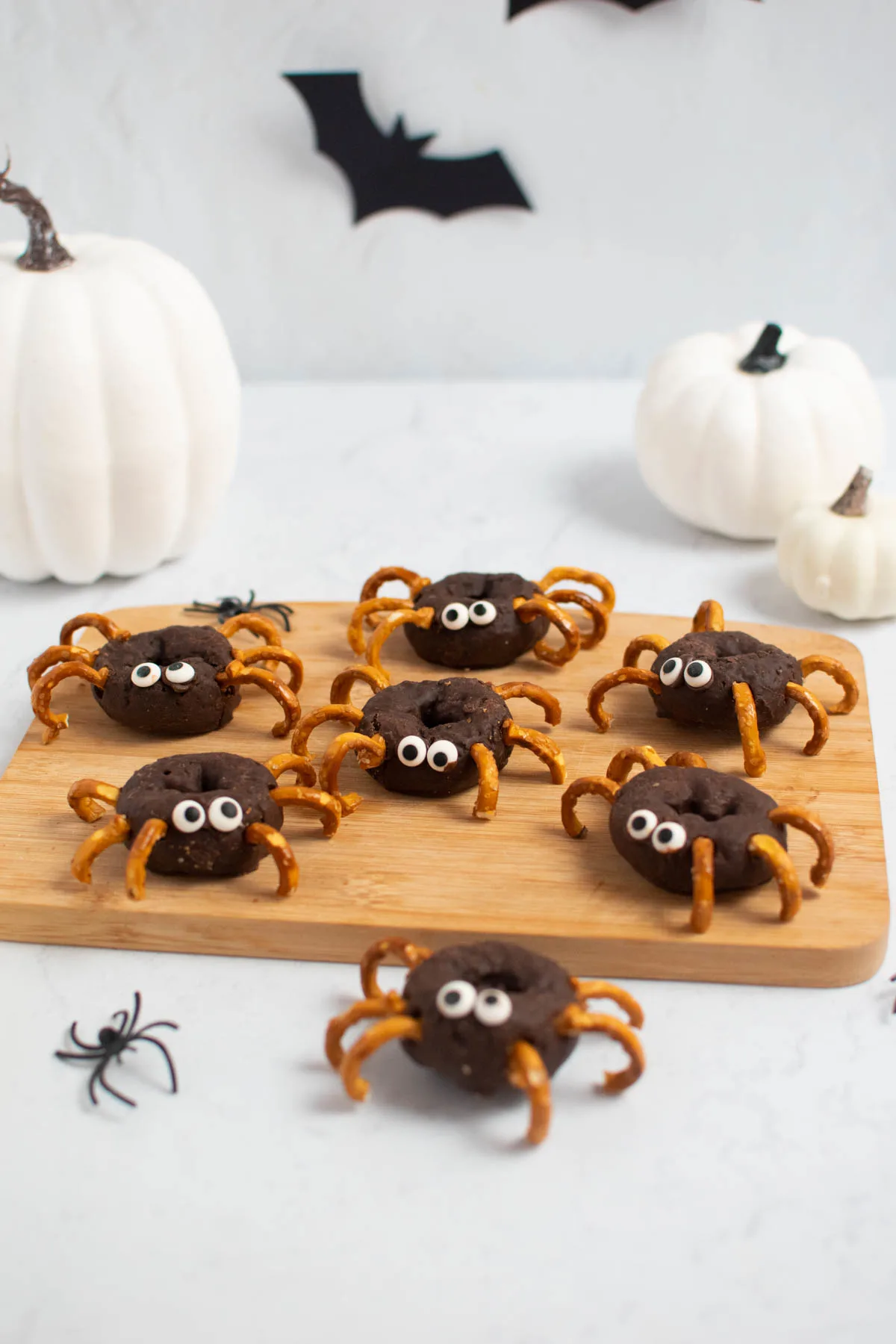 Halloween donut spiders on wood cutting board with white pumpkins and plastic spiders nearby.