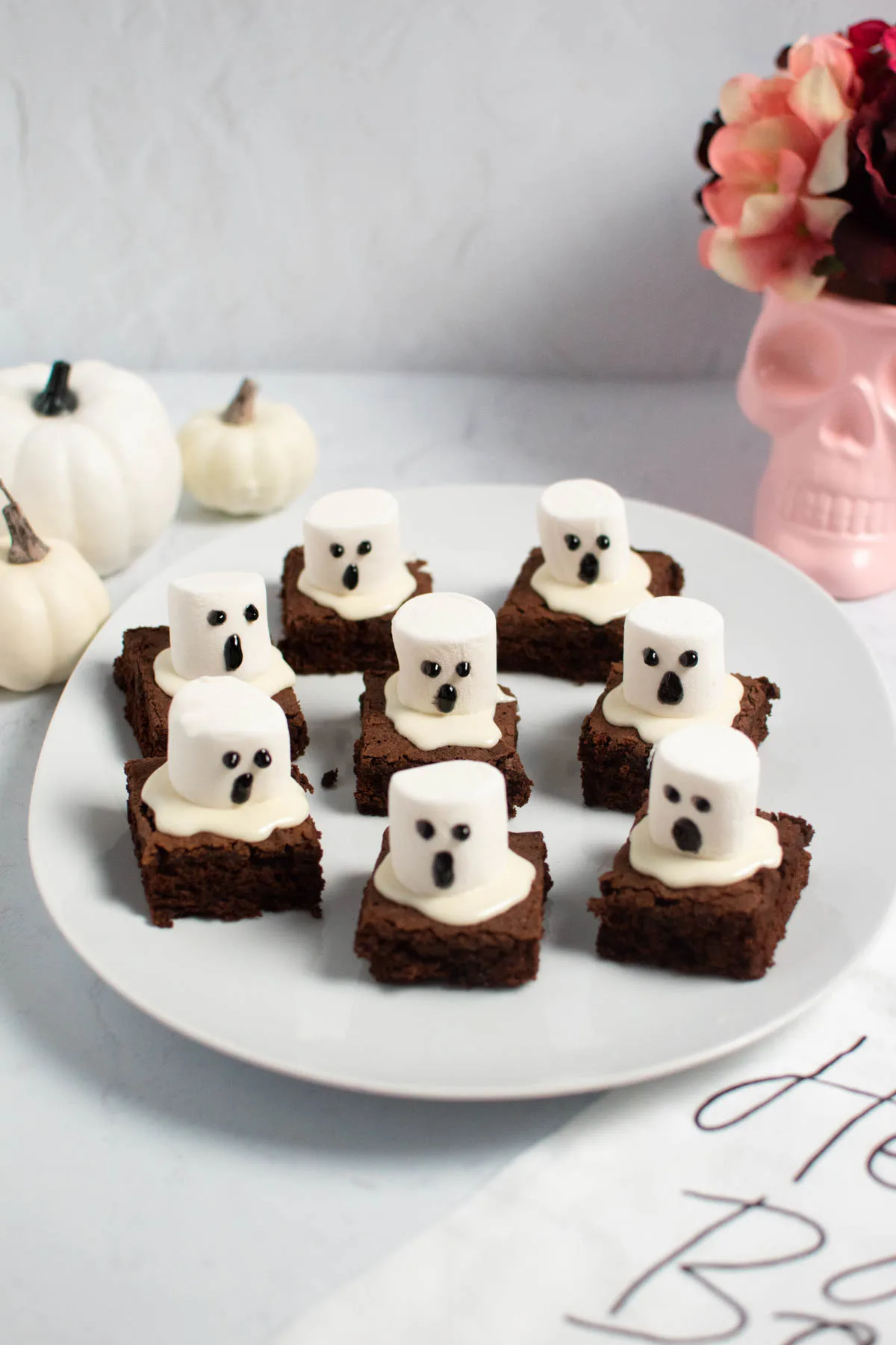 Halloween ghost brownies on white platter with cream pumpkins and pink skull vase in background.