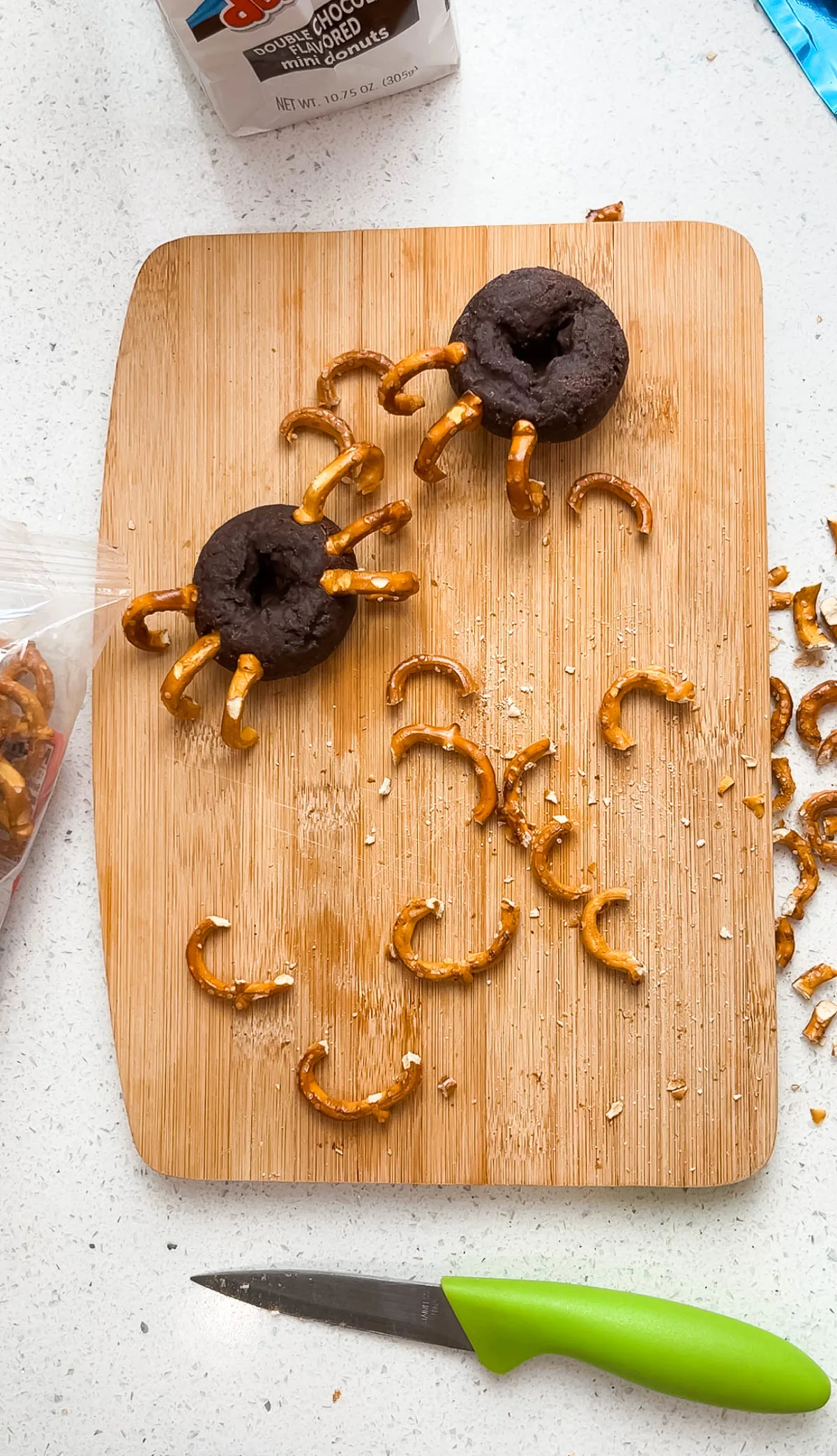 Two mini chocolate donuts with preztel legs surrounded by broken pretzel pieces.