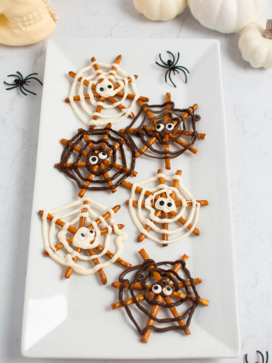 Pretzel spider webs with candy eyes on platter with plastic spiders and pumpkins.