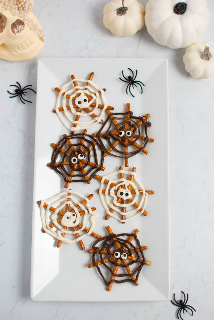 Pretzel stick spider webs with plastic spiders, pumpkins, and skull head nearby.