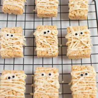 Nine rice krispie mummies with candy eyes on black baking rack.