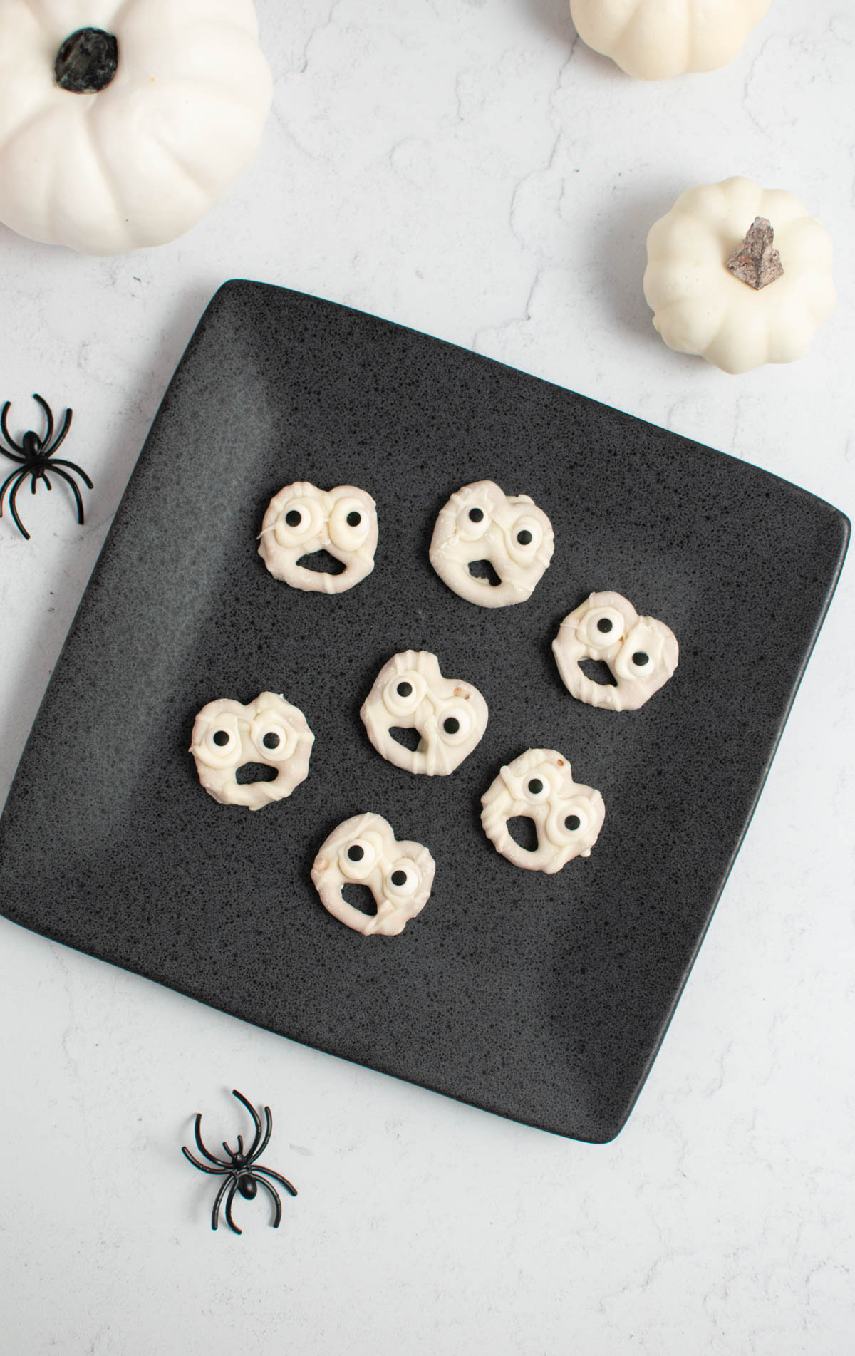 Seven white chocolate ghost pretzels on gray square plate with plastic spiders and white pumpkins nearby.