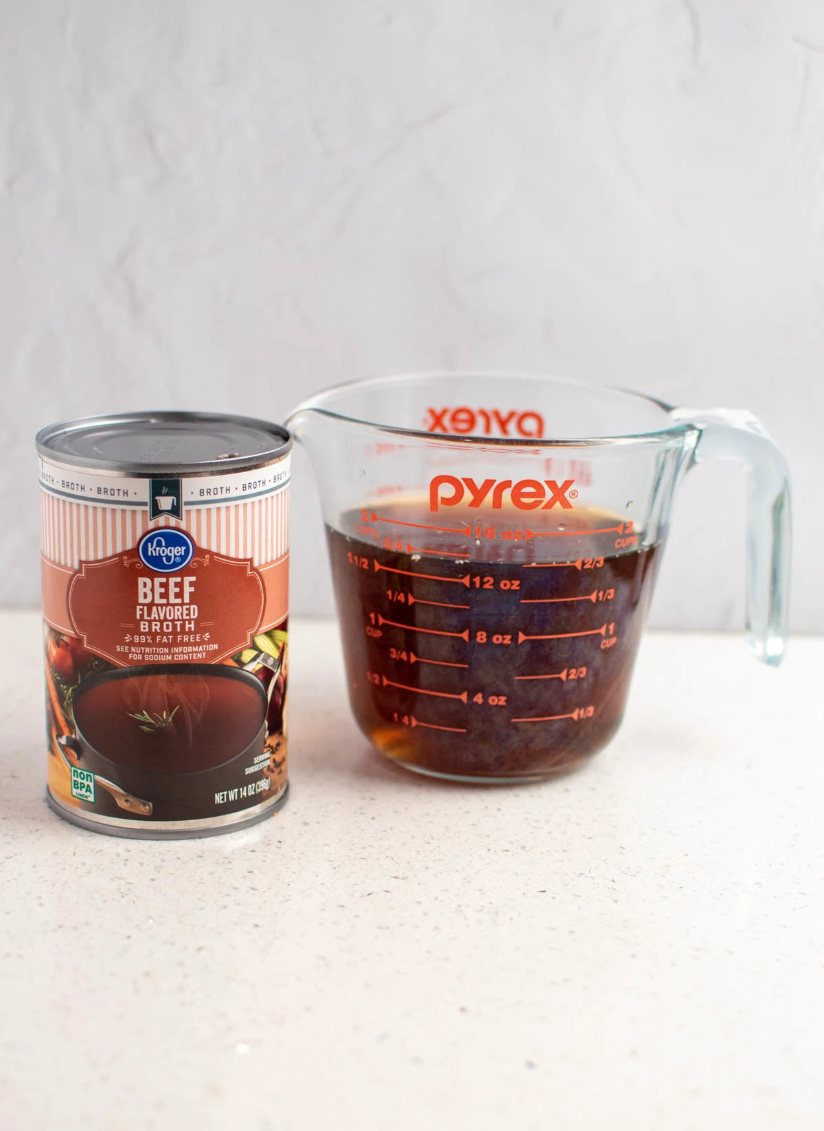 Beef broth in a glass measuring cup next to can of beef broth all on white kitchen counter.